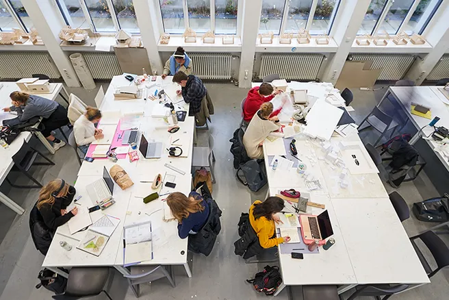 Studierende aus der Vogelperspektive konzentriert am Arbeiten im Zeichensaal. Auf den weißen Arbeitstischen sind Papiere, Zeichnungen und Laptops verteilt, auf der Fensterbank im Hintergrund stehen kleine Modelle aus Holz.