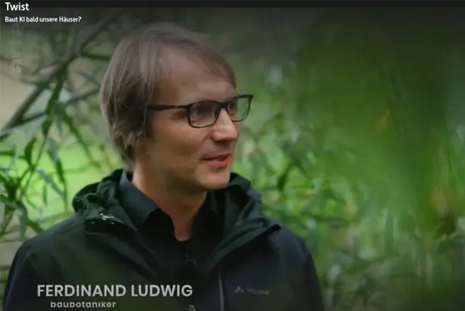 Mann mittleren Alters mit schwarzem Hemd und Jacke vor grünem, pflanzlichen Hintergrund